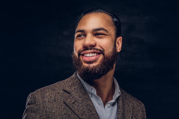 Foto gratuita retrato de primer plano de un hombre de negocios afroamericano barbudo guapo y sonriente con una chaqueta clásica marrón. aislado en un fondo oscuro.