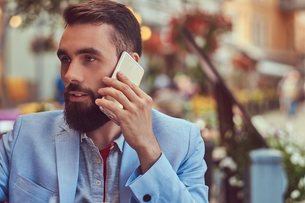 Retrato de primer plano de un hombre de negocios barbudo de moda con un corte de pelo elegante, hablando por teléfono, bebe un vaso de jugo fresco, sentado en un café al aire libre.