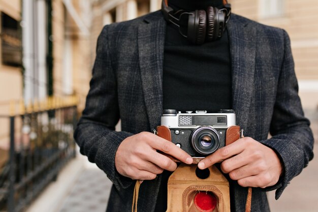 Retrato de primer plano de hombre viste camisa negra y chaqueta gris posando en la calle por la mañana. Foto de fotógrafo con cámara de sujeción de piel marrón claro.