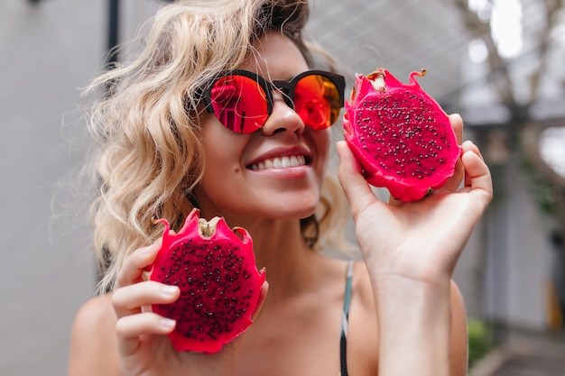 Retrato de primer plano de una impresionante chica rubia con gafas de sol rosas posando con frutas exóticas. Foto de modelo femenino rizado riendo con pitahaya roja.