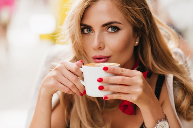 Retrato de primer plano de la impresionante dama de ojos azules relajándose en la cafetería y bebiendo café caliente