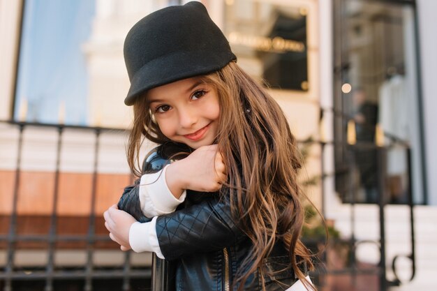 Retrato de primer plano de un increíble niño de pelo largo con una maravillosa sonrisa amable posando afuera, abrazando el pilar de hierro.