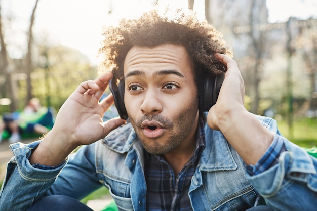 Retrato de primer plano de joven afroamericano emocionado y fascinado con peinado afro, sentado en partk y escuchando música en auriculares mientras los sostiene con las manos.