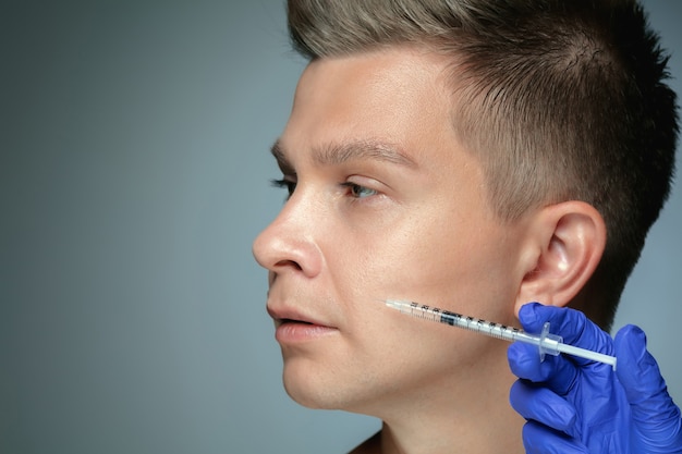 Retrato de primer plano de joven aislado en pared gris. Procedimiento de cirugía de relleno, labios y pómulos. Concepto de salud y belleza masculina, cosmetología, cuerpo y cuidado de la piel. Anti-envejecimiento.