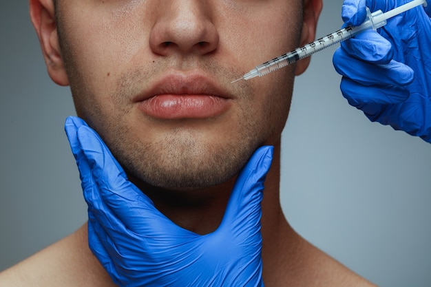 Retrato de primer plano de joven aislado sobre fondo gris de estudio. Procedimiento de cirugía de llenado. Concepto de salud y belleza masculina, cosmetología, autocuidado, cuidado corporal y de la piel. Anti-envejecimiento.