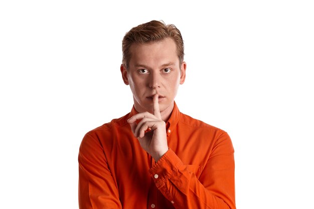 Retrato de primer plano de un joven apuesto jengibre con una elegante camisa naranja mirando a la cámara y mostrando un signo silencioso mientras posa aislado en el fondo blanco del estudio. Expresiones faciales humanas. Sinc