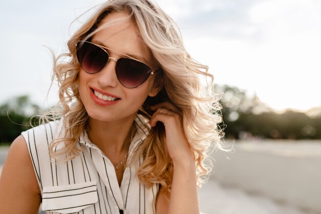 Retrato de primer plano de la joven y atractiva mujer rubia elegante en las calles de la ciudad en vestido de estilo de moda de verano con gafas de sol