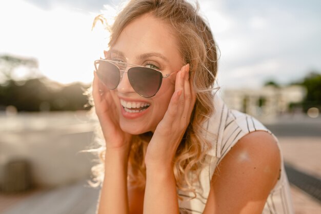 Retrato de primer plano de la joven y atractiva mujer rubia con estilo en las calles de la ciudad en vestido de estilo de moda de verano con gafas de sol