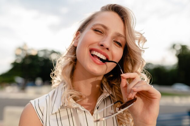 Retrato de primer plano de la joven y atractiva mujer rubia con estilo en las calles de la ciudad en vestido de estilo de moda de verano con gafas de sol