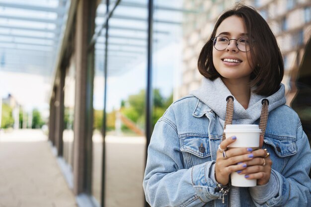 Retrato de primer plano de una joven bonita y feliz que explora la ciudad y mira hacia otro lado con una cara alegre y complacida bebiendo una taza de café para llevar de la calle peatonal local favorita al aire libre disfrutando de la primavera