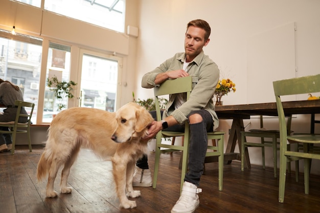 Foto gratuita retrato en primer plano de un joven guapo esperando su pedido en una cafetería acariciando a un perro descansando