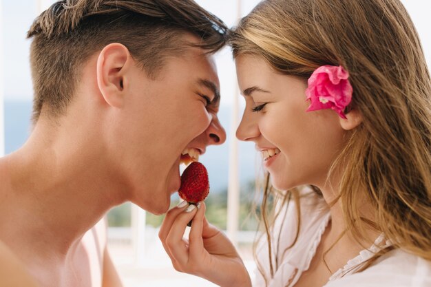 Retrato de primer plano de la joven pareja amorosa posando con los ojos cerrados, mientras come fresa. Adorable chica rubia con flor rosa en el pelo alimenta a su novio con sabrosas bayas.