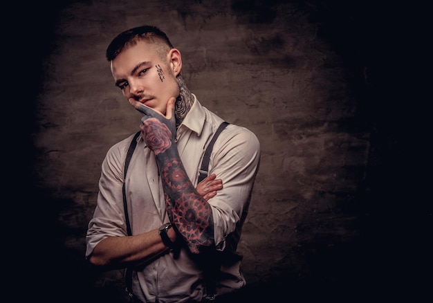 Retrato de primer plano de un joven pensativo tatuado a la antigua usanza con camisa blanca y tirantes sostiene la mano en la barbilla. Aislado en un fondo oscuro.