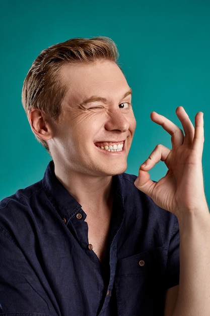 Foto gratuita retrato de primer plano de un joven peson de jengibre con una elegante camiseta azul marino que muestra el signo de ok, mirando a la cámara y sonriendo mientras posa en el fondo azul del estudio. expresiones faciales humanas. sincero