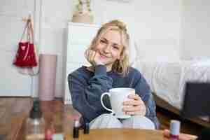 Foto gratuita retrato en primer plano de una joven rubia sentada en su habitación mirando a la cámara sosteniendo una taza de té