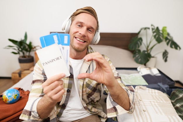 Foto gratuita retrato en primer plano de un joven turista guapo y feliz que muestra un pasaporte y boletos de avión con signo de corazón