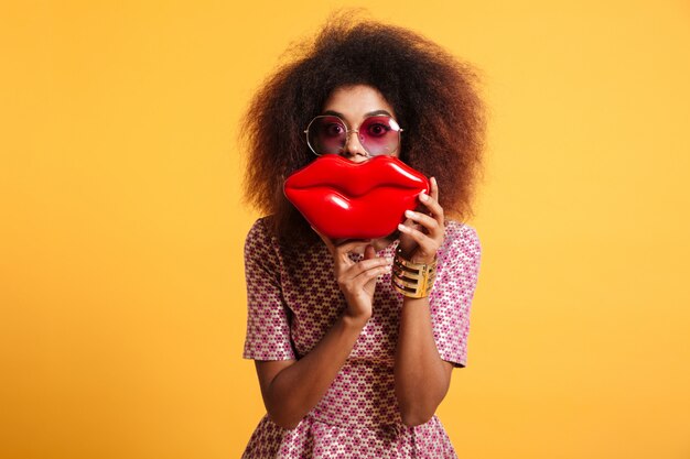 Retrato de primer plano de juguetón wooman africano en gafas de sol con grandes labios rojos delante de su cara