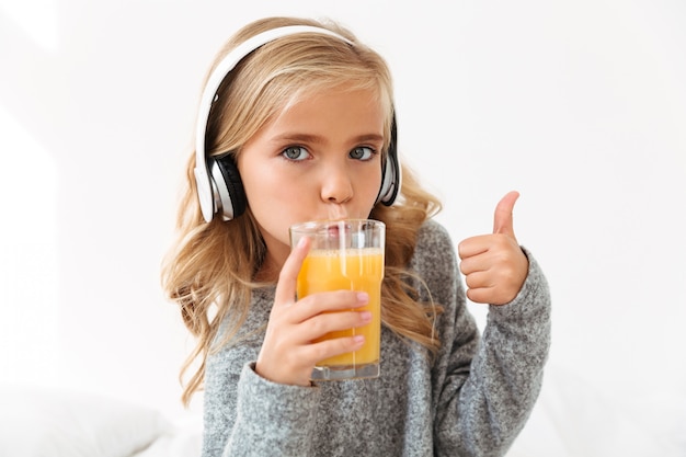 Retrato de primer plano de linda chica en auriculares bebiendo jugo de naranja, mostrando el pulgar hacia arriba gesto