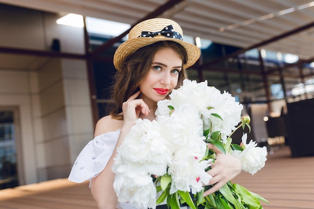 Retrato de primer plano de linda chica con pelo largo con sombrero está sentado en el suelo en la terraza. Lleva un vestido blanco con hombros desnudos, lápiz labial rojo. Ella tiene flores blancas en las manos y sonríe.