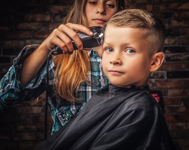 Retrato de primer plano de un lindo niño en edad preescolar que se corta el pelo. La hermana mayor corta a su hermano pequeño con una podadora contra una pared de ladrillos.
