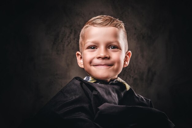 Retrato de primer plano de un lindo niño sonriente sentado en una silla con la capa negra del salón y espera al peluquero.