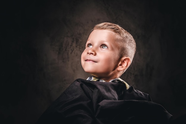 Retrato de primer plano de un lindo niño sonriente sentado en una silla con la capa negra del salón y espera a la peluquería.