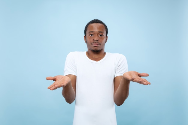 Retrato de primer plano de medio cuerpo del joven modelo masculino afroamericano en camisa blanca en el espacio azul. Las emociones humanas, la expresión facial, el concepto de publicidad.