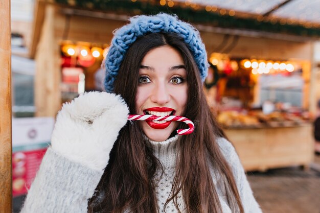 Retrato de primer plano de modelo femenino divertido con cabello oscuro comiendo con placer bastón de caramelo en Navidad. Chica morena alegre en guantes blancos disfrutando de piruleta en día frío.