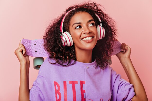 Retrato de primer plano de modelo femenino positivo en camisa púrpura mirando hacia arriba con una sonrisa. Hermosa joven africana escuchando su canción favorita en auriculares.