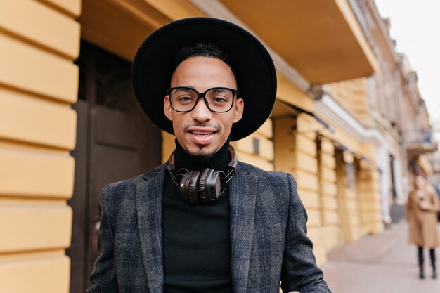 Retrato de primer plano del modelo masculino africano cansado con grandes auriculares de pie cerca del restaurante. Foto al aire libre de apuesto hombre seguro de piel oscura.