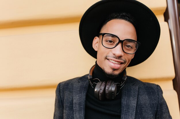 Retrato de primer plano de modelo masculino con piel oscura que expresa emociones positivas. Foto al aire libre del adorable hombre negro caminando con auriculares en fin de semana.