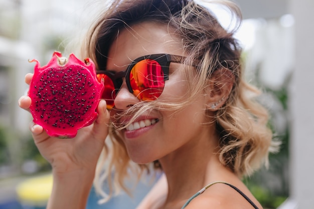 Foto gratuita retrato de primer plano de la modelo de mujer caucásica alegre lleva gafas rosas durante la sesión de fotos en el resort. sonriente mujer blanca con fruta de dragón rojo