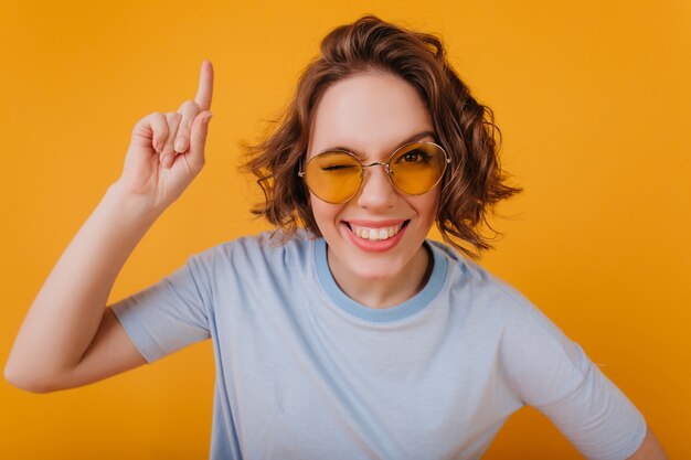 Retrato de primer plano de mujer adorable con sonrisa sincera divirtiéndose en estudio amarillo