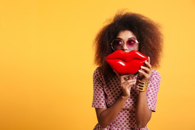 Retrato de primer plano de mujer afroamericana divertida en gafas de sol con grandes labios rojos frente a su cara, mirando a un lado