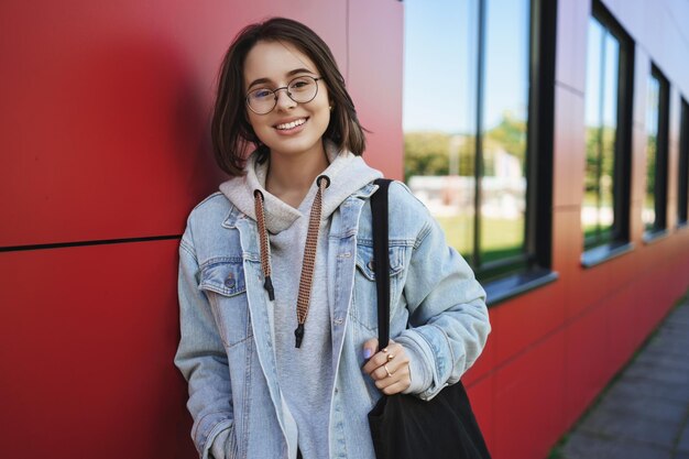 Retrato de primer plano de una mujer alegre y bastante joven de 20 años con gafas, estudiante o empleado, cámara sonriente, feliz llevando una bolsa de asas, pared de edificio de ladrillo rojo magra del campus disfrutando del ocio