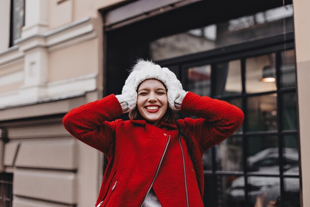 Retrato de primer plano de mujer alegre con lápiz labial rojo, riendo con los ojos cerrados. Chica con abrigo, gorro y guantes toca su cabeza.