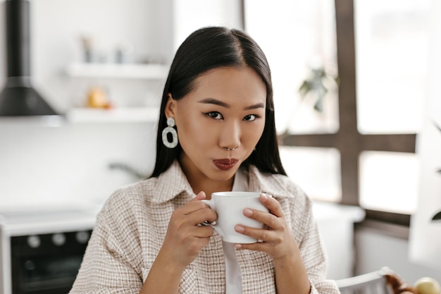 Retrato de primer plano de una mujer asiática morena bronceada con lápiz labial oscuro bebe té Lady mira a la cámara y sostiene una taza de café con leche en la cocina