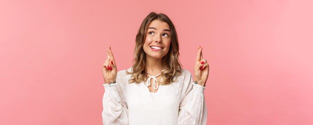 Retrato de primer plano de una mujer atractiva, optimista y esperanzada con vestido blanco que muerde el labio y sonríe mientras sueña despierto, el sueño de esperanza se hace realidad, pide un deseo o reza de pie con fondo rosa