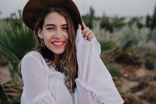 Retrato de primer plano de mujer atractiva con sombrero marrón y blusa blanca posando con linda sonrisa. Hermosa mujer joven sin maquillaje pasar tiempo afuera disfruta de la noche y la vista de la naturaleza