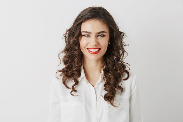 Retrato de primer plano de una mujer atractiva sonriente con dientes blancos, pelo largo y rizado, maquillaje de lápiz labial rojo mirando en cámara aislada con blusa blanca