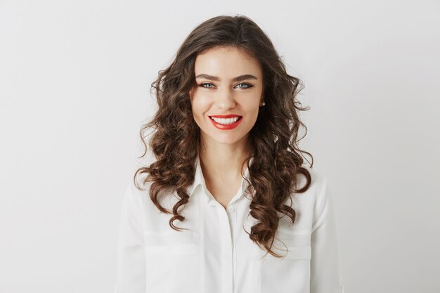 Retrato de primer plano de una mujer atractiva sonriente con dientes blancos, pelo largo y rizado, maquillaje de lápiz labial rojo mirando en cámara aislada con blusa blanca