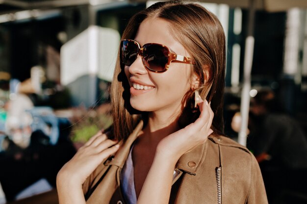 Retrato de primer plano de mujer blanca alegre con gafas tocando su cabello sobre fondo borroso. Señora bonita con sonrisa maravillosa pasar tiempo al aire libre y muestra buen humor