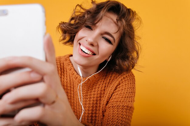 Retrato de primer plano de mujer blanca complacida con maquillaje casual tomando una foto con su teléfono