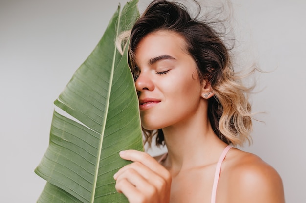 Retrato de primer plano de mujer caucásica alegre sin maquillaje posando. Foto interior de una chica bastante rubia con hojas verdes aisladas.