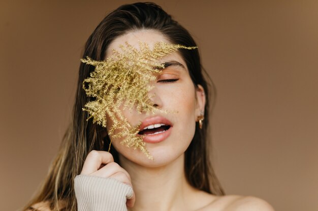 Retrato de primer plano de mujer europea alegre con planta. Hermosa modelo de mujer morena posando en la pared marrón con la boca abierta.