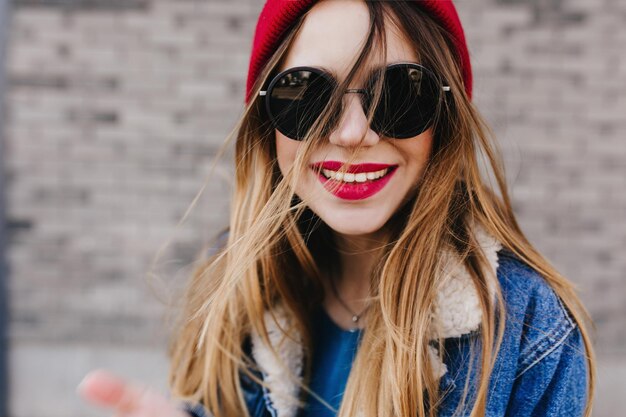 Retrato de primer plano de mujer europea con sombrero rojo sobre fondo urbano borroso Chica hermosa riendo con gafas de sol negras posando cerca de la pared de ladrillo