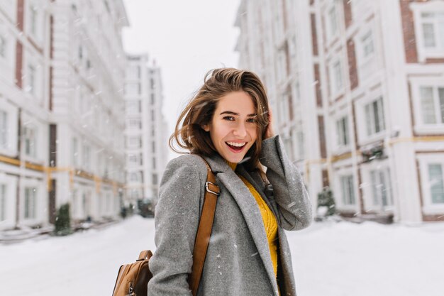 Retrato de primer plano de mujer extática en elegante abrigo gris de pie en la calle en día de nieve. Foto al aire libre del modelo femenino de moda con bolso marrón caminando por la ciudad en vacaciones de invierno.