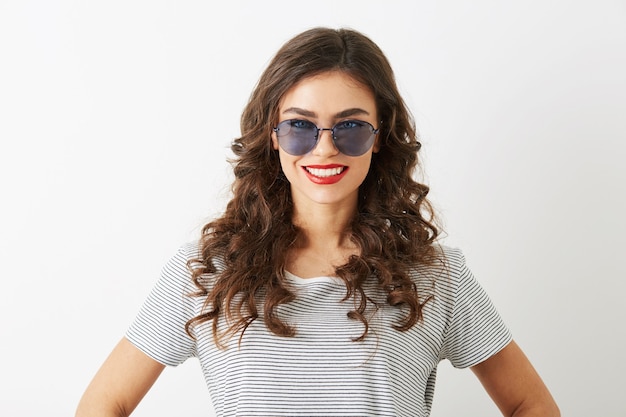 Retrato de primer plano de una mujer joven atractiva con el pelo rizado sonriendo aislado sobre fondo blanco con gafas de sol vestidas con camiseta