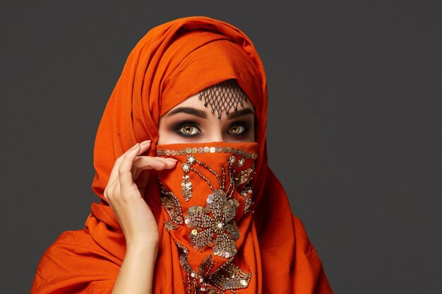 Retrato de primer plano de una mujer joven de buen aspecto con hermosos ojos ahumados y joyas finas en la frente, vistiendo el hiyab de terracota decorado con lentejuelas. Ella está posando de lado y mirando t