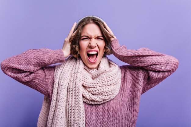 Retrato de primer plano de mujer joven enojada en bufanda tejida. Foto interior de niña emocional de pelo corto posando con expresión de cara molesta.